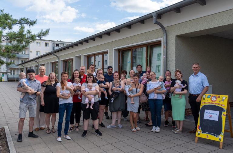Eine Gruppe von Menschen mit Baby in ihren Händen stehen vor einem Gebäude und schauen in die Kamera.