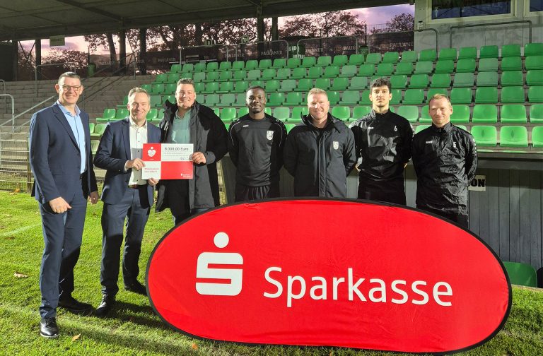 Eine Gruppe von 7 Männern stehen in einem Fußballstadion hinter einem roten Sparkassen-Banner
