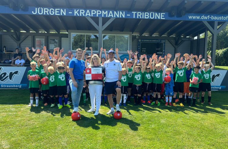 Die Mannschaft von Germania 90 Schöneiche steht im Hintergrund in ihrem Fußballstadion. Vor ihnen stehen drei Personen, die einen Scheck in die Kamera halten.