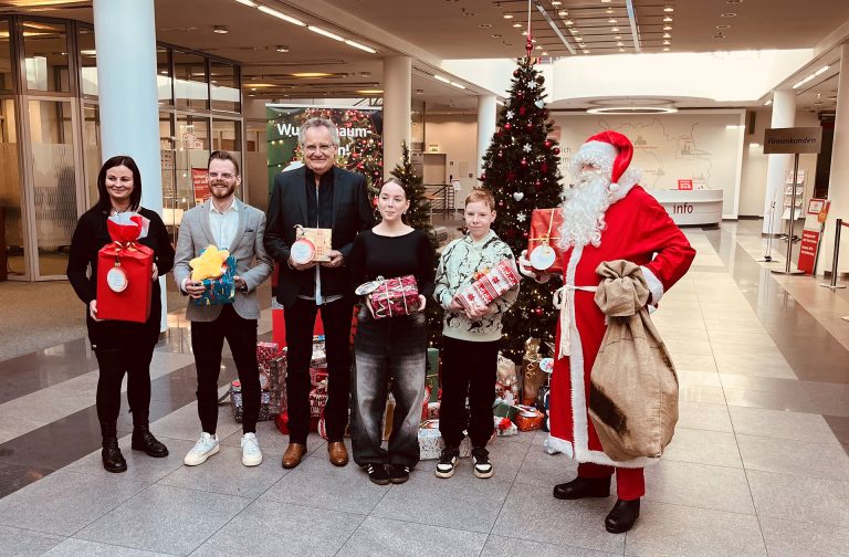 6 Menschen, darunter auf der rechten Seite ein als Weihnachtsmann verkleideter Mann, stehen vor einem Weihnachtsbaum.