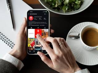 Das Bild ist von der Vogelperspektive aufgenommen. Man sieht zwei Hände, die ein Smartphone halten. Auf dem Smartphone ist eine Website der Sparkasse zu sehen. Rechts neben den Händen sieht man eine Kaffeetasse mit Kaffee darin.