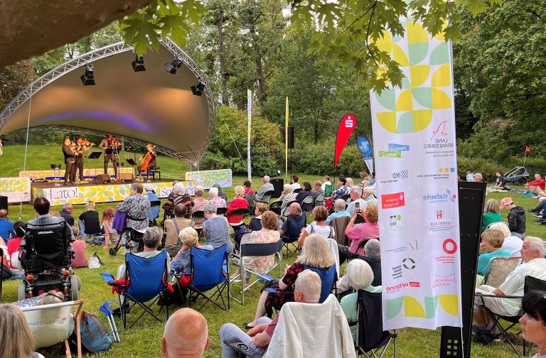 Im Foto ist auf der linken Seite im Hintergrund eine Bühne mit drei Geigern und einem Cellisten zu sehen, die gerade spielen. Vor ihnen sitzen auf Stühlen oder im Gras Menschen, die sich das Konzert anhören. Das alles spielt in einem grünen Park.